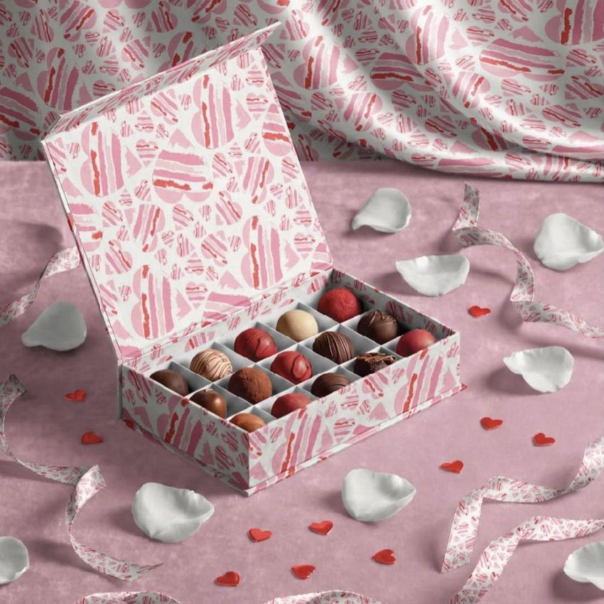Open chocolate box mockup with assorted pralines displayed on a pink surface, surrounded by white rose petals and small red hearts.