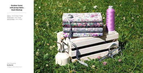 Stack of folded jersey fabrics with floral patterns on a wooden crate, surrounded by sewing tools and thread, outdoors on grass.