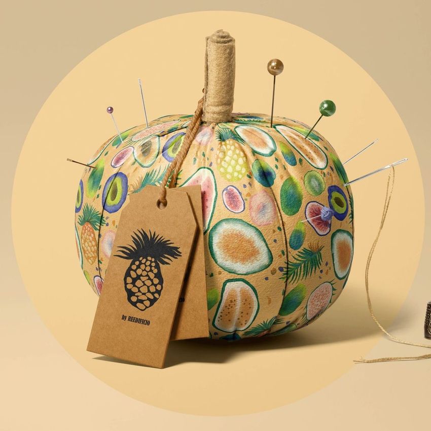 Fabric pumpkin mockup with a colorful tropical fruit pattern, pins inserted on top, and a brown tag with a pineapple graphic.
