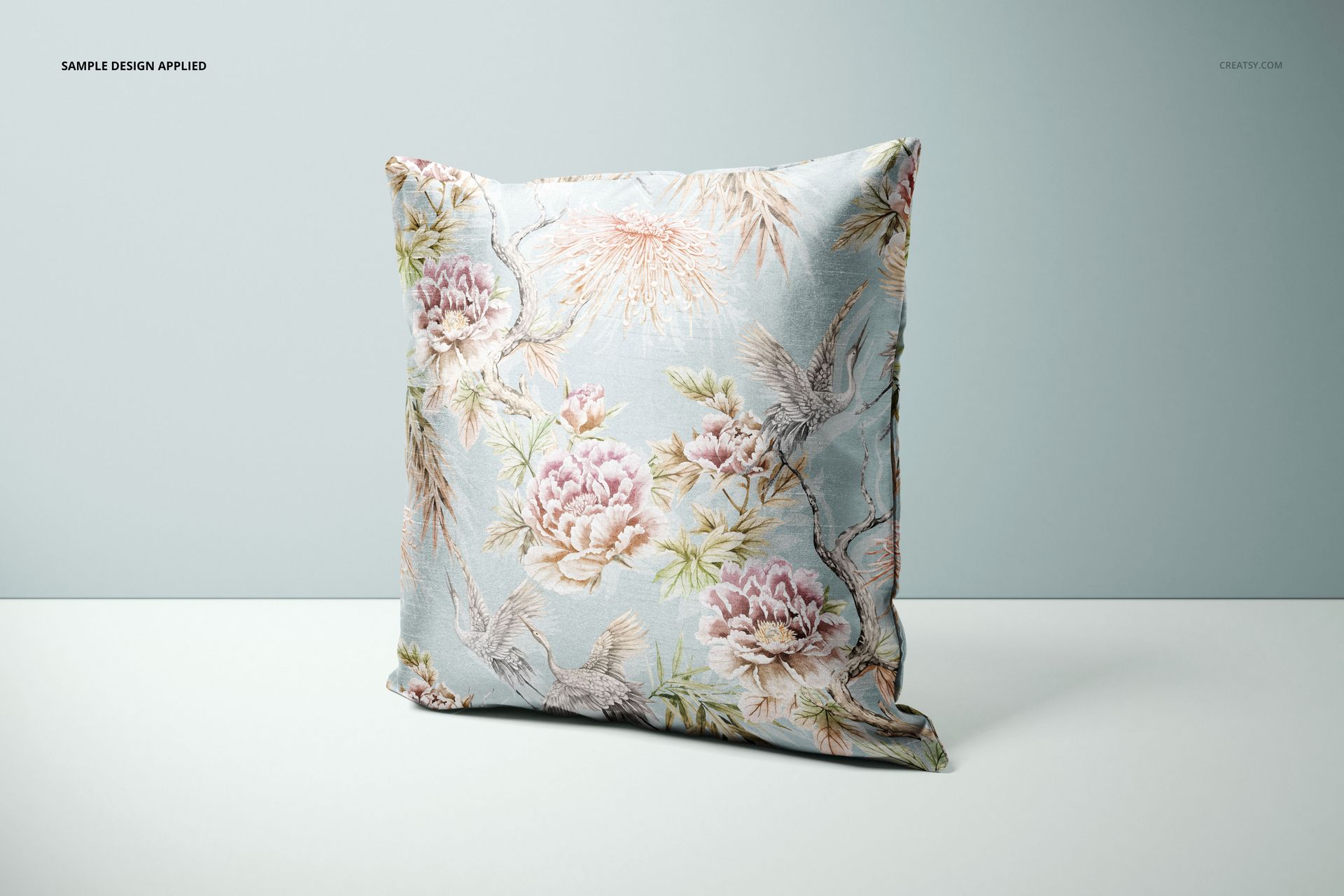 Front-facing cushion cover mockup featuring pastel floral and crane motifs on a soft blue background, minimal scene.