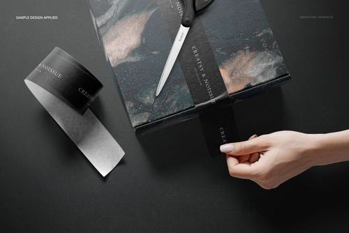 Mailing box with dark marbled pattern on a black surface, hand applying black branded tape, scissors nearby.