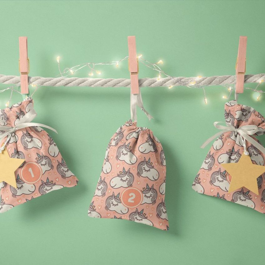 Drawstring sacks advent calendar mockup with three pink fabric bags featuring hand-drawn unicorn patterns, hanging on a rope with clothespins against a pastel green background.