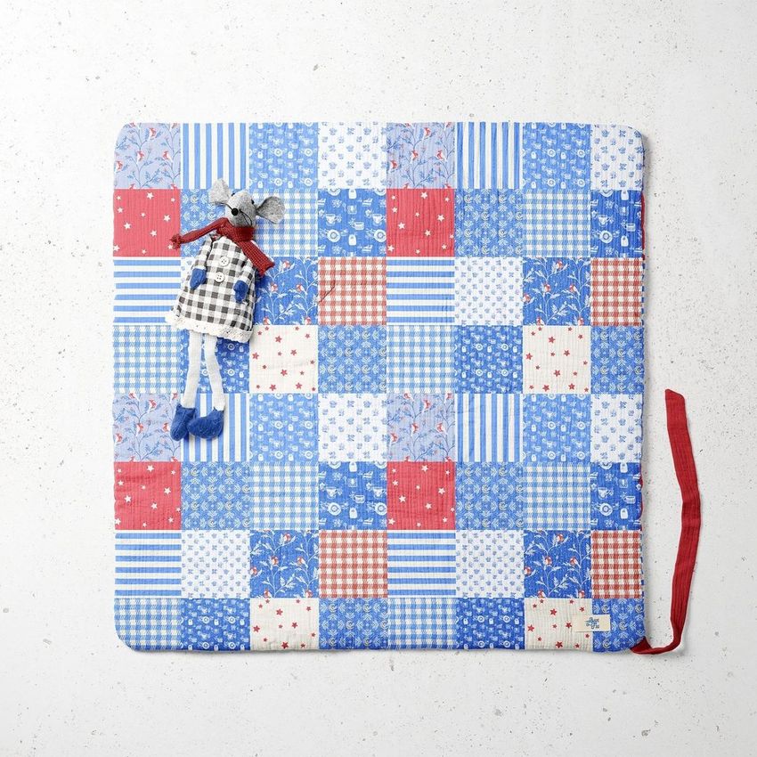 Top-down view of a newborn swaddle wrap mockup with a blue, red, and white patchwork pattern and a small fabric mouse toy on top.