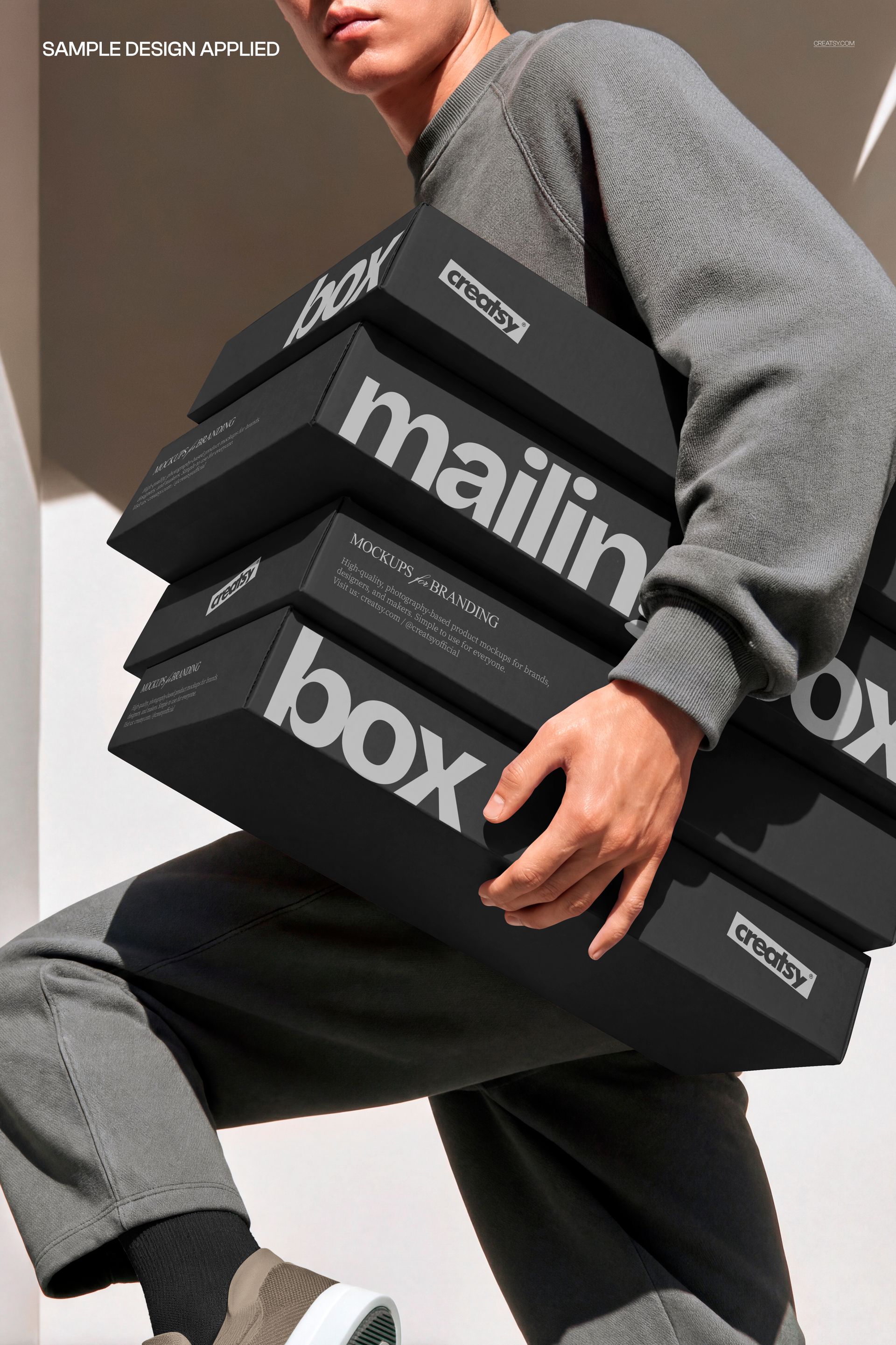 Person in casual gray clothing holding a stack of black mailing boxes with bold white text, front view mockup.