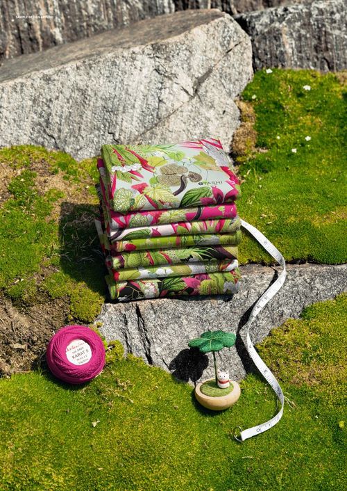 PSD mockup showing folded fabrics with botanical designs on moss-covered rocks, featuring sewing accessories in the scene.