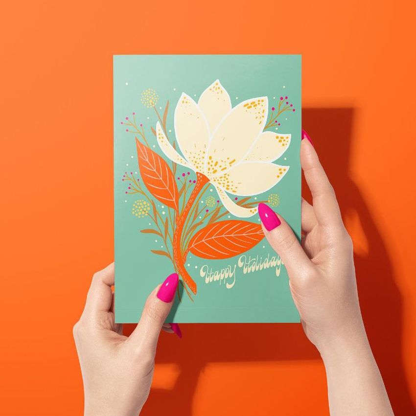 Greeting card mockup featuring a woman's hands holding a card with a large white magnolia flower and orange leaves on a teal background.