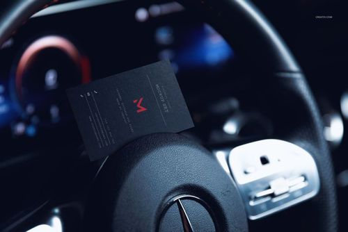 Black automotive business card mockup resting on a car steering wheel, close-up view with dashboard in background.