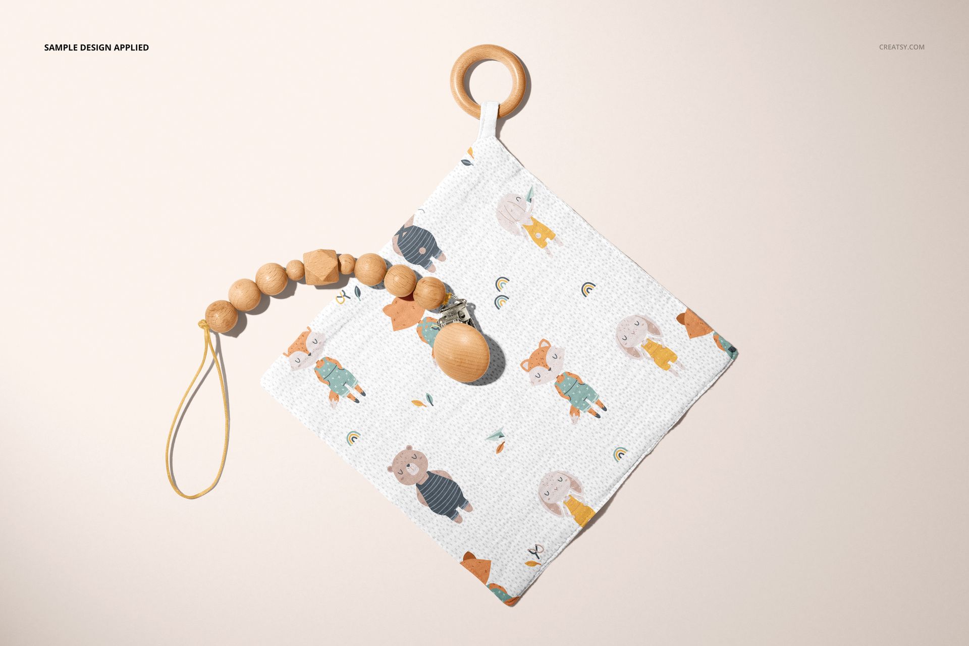 PSD mockup of a muslin swaddle with playful animal patterns and a wooden teether, shown flat against a beige backdrop.