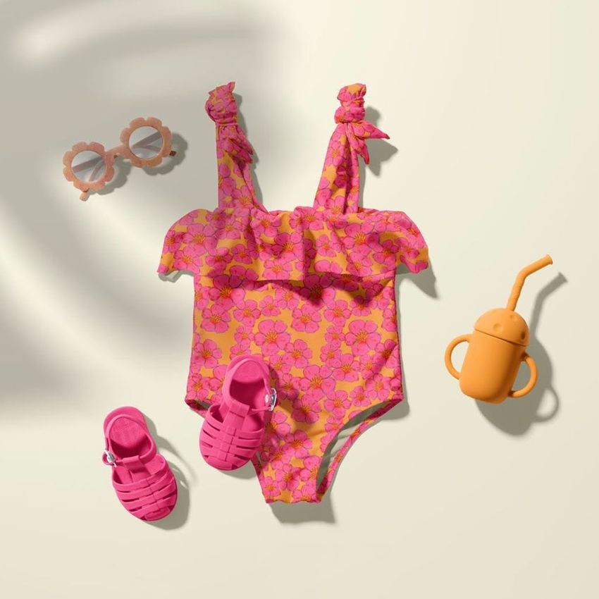 Baby swimsuit mockup featuring a pink floral one-piece laid flat with matching pink sandals, sunglasses, and an orange sippy cup on a light background.