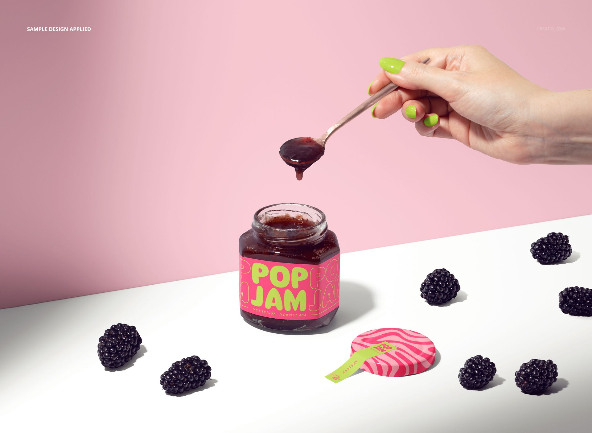 Front view of a jam jar mockup featuring a colorful label, loose blackberries, and a spoon held by a hand.
