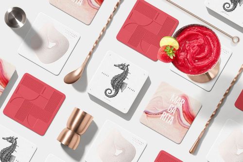 Square coasters arranged on a white surface with cocktail tools, featuring red and white designs and a drink garnish.