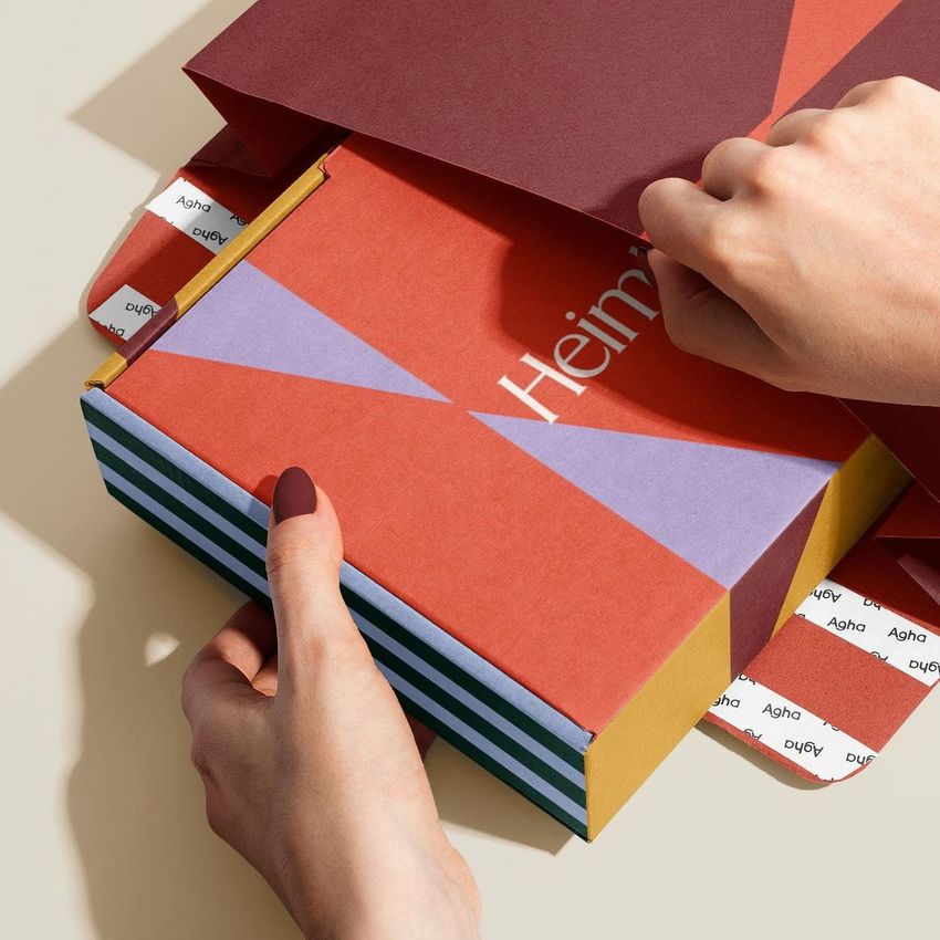 Mailer box mockup with bold geometric patterns in red, purple, and yellow, held by hands on a neutral background.