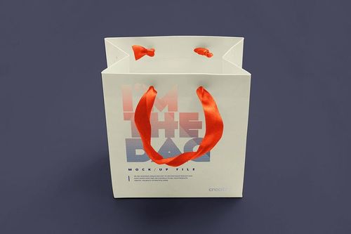 Square-shaped gift bag mockup shown from above, featuring red handles and printed text on a white background.