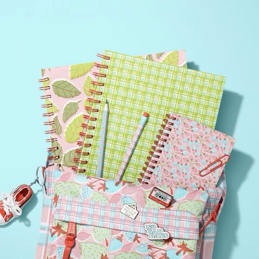 School backpack mockup with three spiral notebooks in pastel patterns, two pencils, and a small sneaker keychain on a blue background.