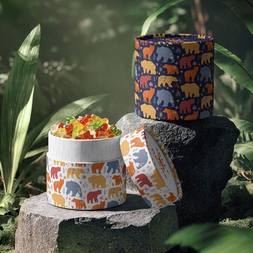 Paper tubes packaging mockup with colorful jungle animal patterns, displayed outdoors on rocks with green foliage and gummy candies.