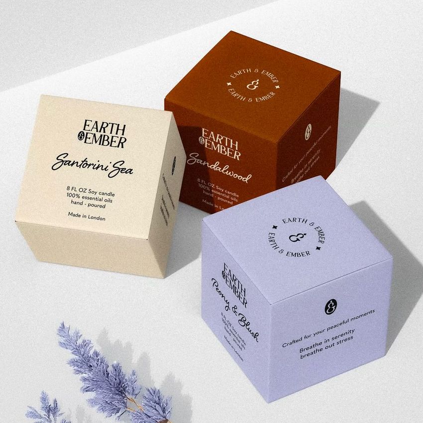 Square box mockup featuring three candle packaging boxes in beige, brown, and lavender on a light surface with a shadowed sprig.