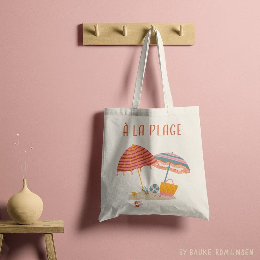 Tote bag mockup hanging on a wooden peg rack against a pink wall, featuring a beach-themed illustration with umbrellas and accessories.