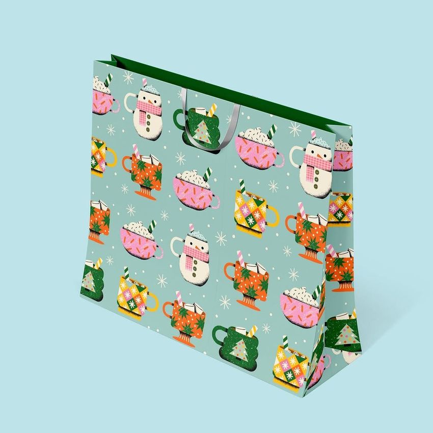 Gift bag mockup with a colorful hot cocoa mug pattern, featuring festive cups and whipped cream on a light blue background.