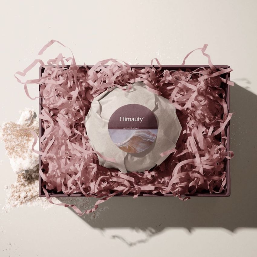 Bath bomb mockup featuring a round wrapped product with a centered label in a box filled with pink shredded paper, viewed from above.