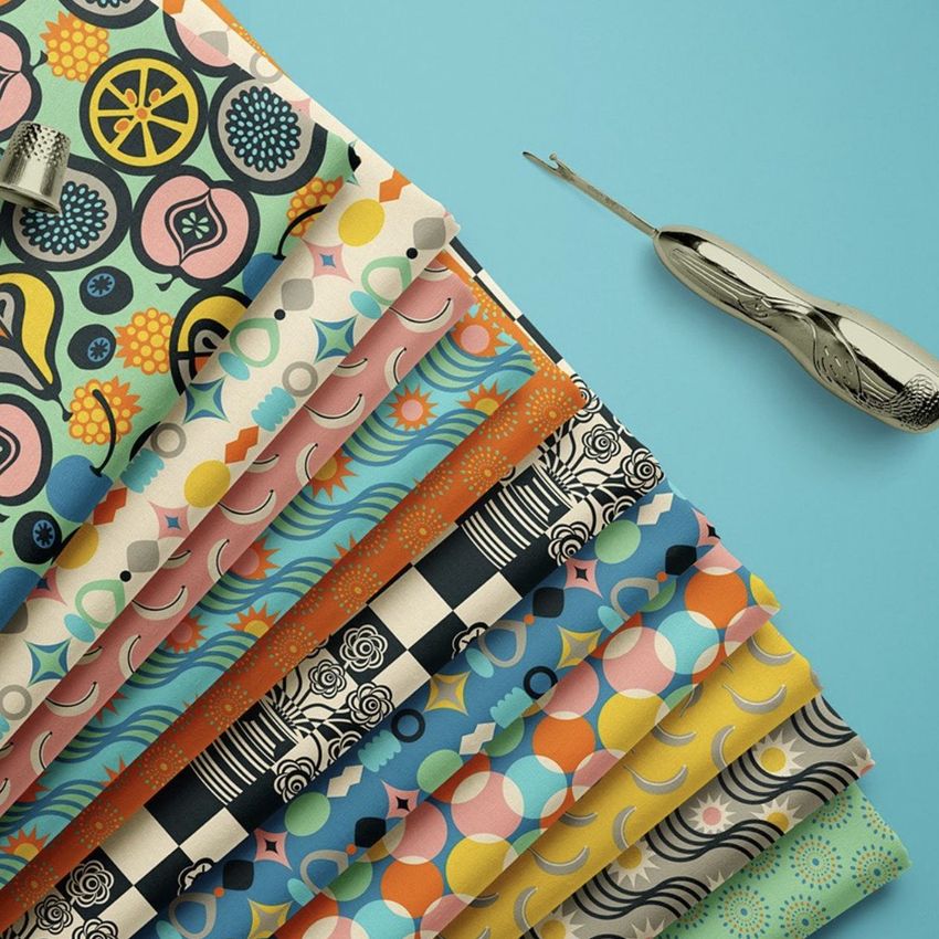 Folded fabrics mockup showing a stack of colorful patterned textiles on a blue surface with a metal seam ripper nearby.