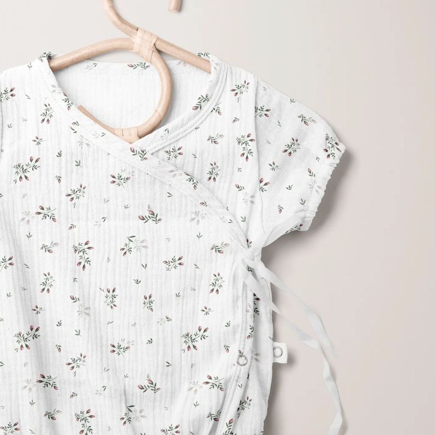 Muslin onesie mockup featuring a short-sleeve baby garment with a delicate floral pattern on a light background, hanging on a wooden hanger.