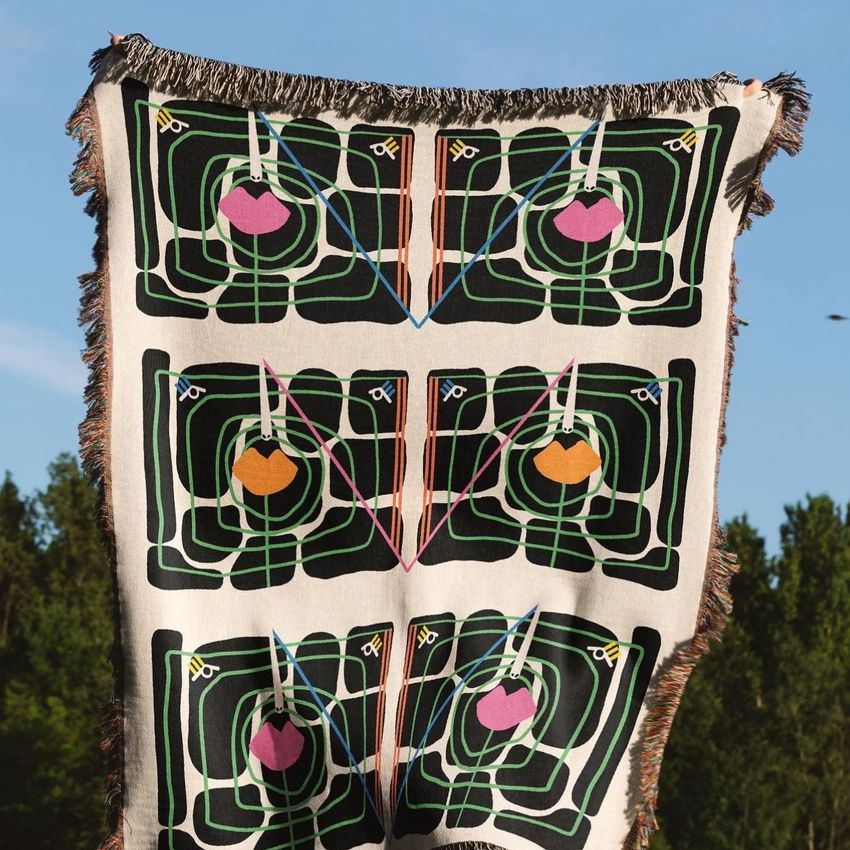 Woven throw blanket mockup held outdoors, featuring an abstract geometric pattern in green, pink, orange, and black tones.