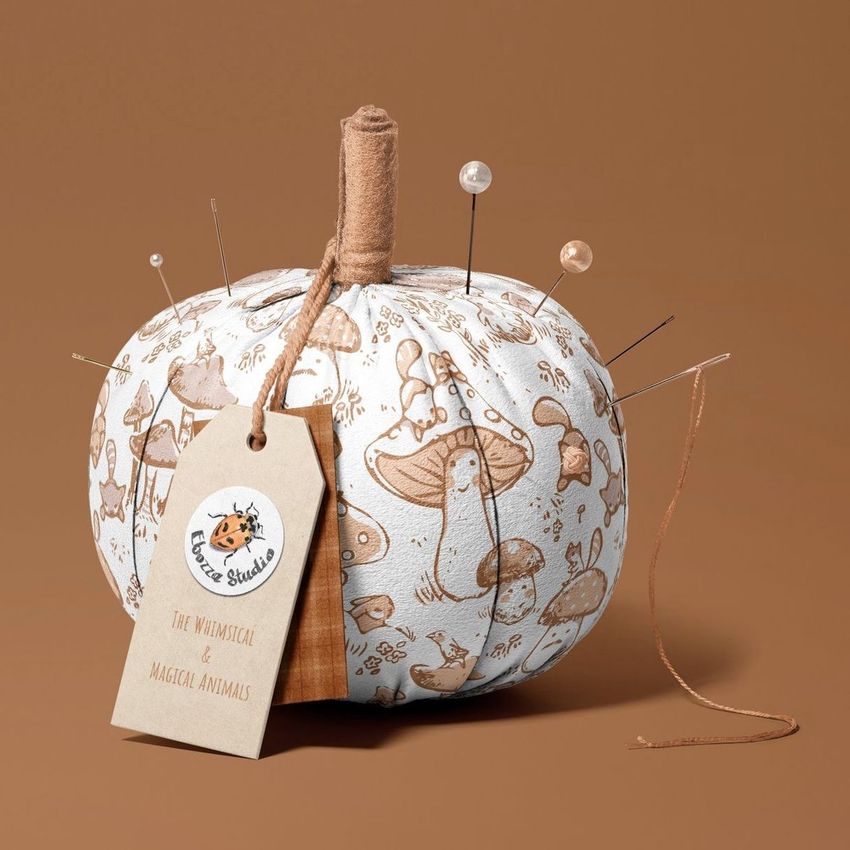 Fabric pumpkin mockup with whimsical animal and mushroom illustrations in brown tones, used as a pincushion with pins and a tag.