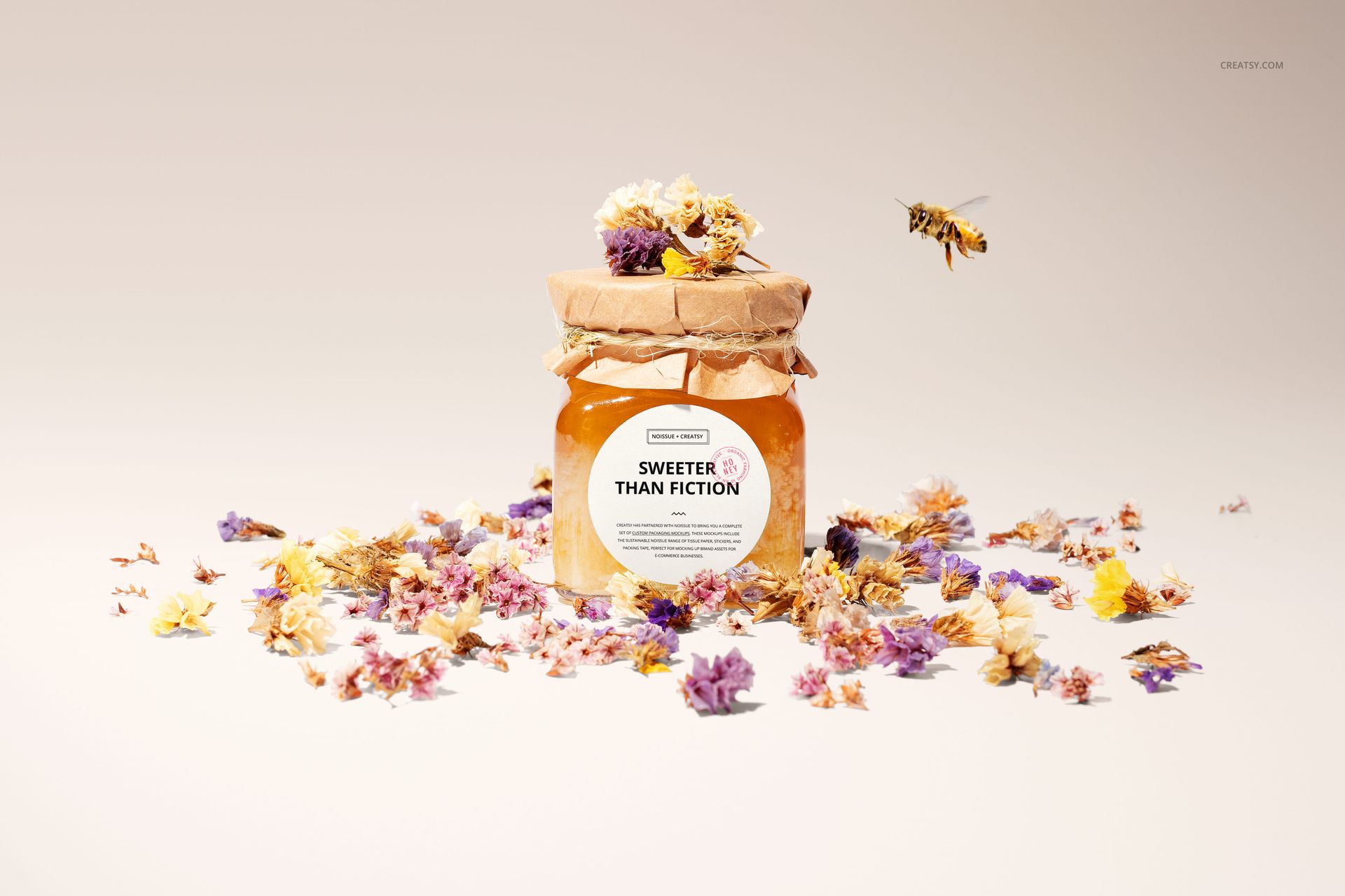 Front view of a flower honey jar mockup with a kraft paper lid, scattered dried flowers, and a bee hovering nearby.