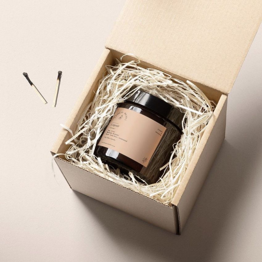 Packaging mockup showing a cardboard box with shredded paper filling and a brown jar inside, viewed from above on a light surface.