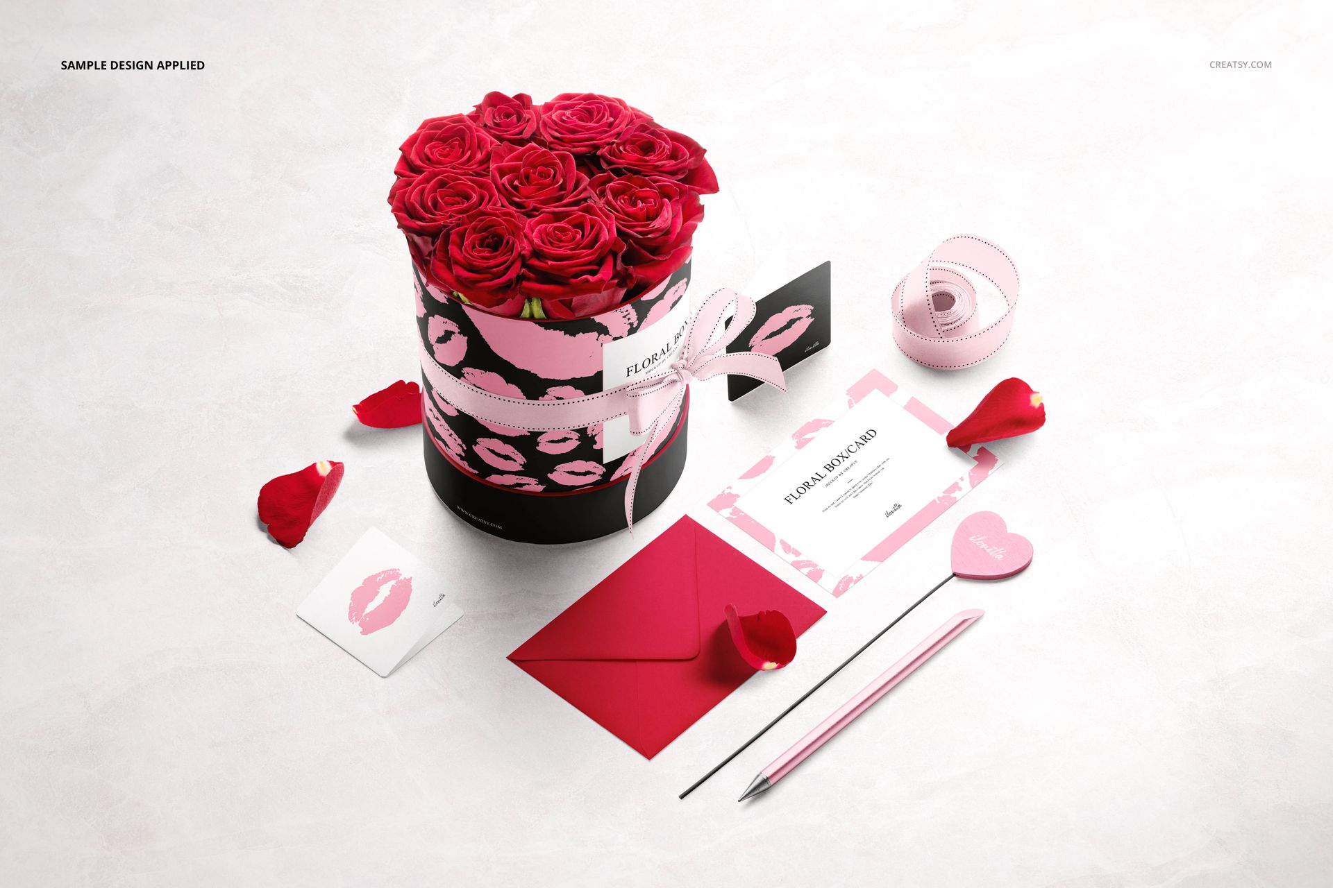 Round floral box mockup with pink roses, surrounded by stationery items, ribbon, and rose petals on a light surface.