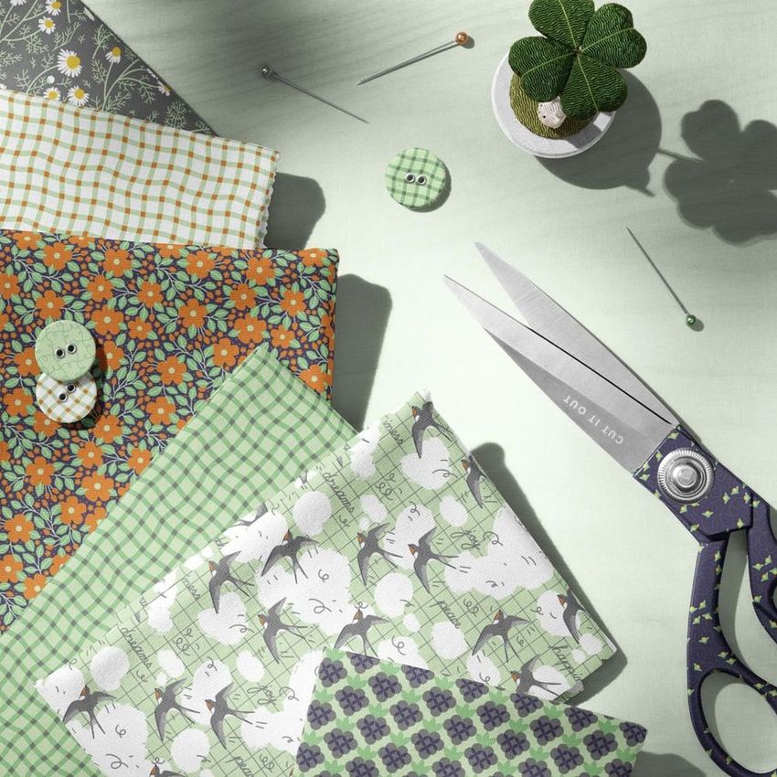 Folded jersey fabrics mockup featuring various patterned textiles in green, orange, and gray, with sewing supplies on a light surface.