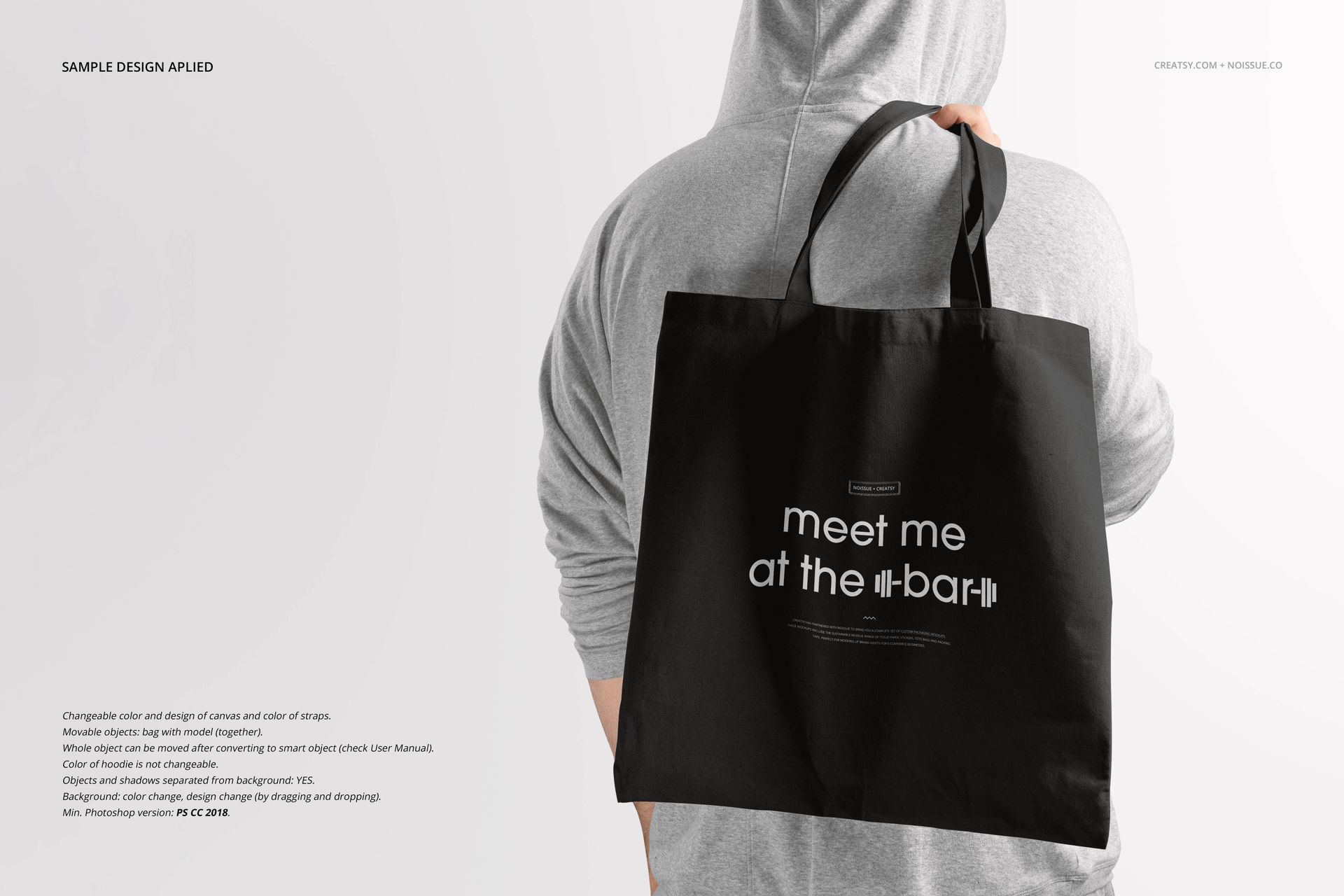 Black tote bag mockup shown from the back, held by a person in a gray hoodie, with white text design on the bag.