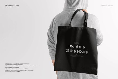 Black tote bag mockup shown from the back, held by a person in a gray hoodie, with white text design on the bag.