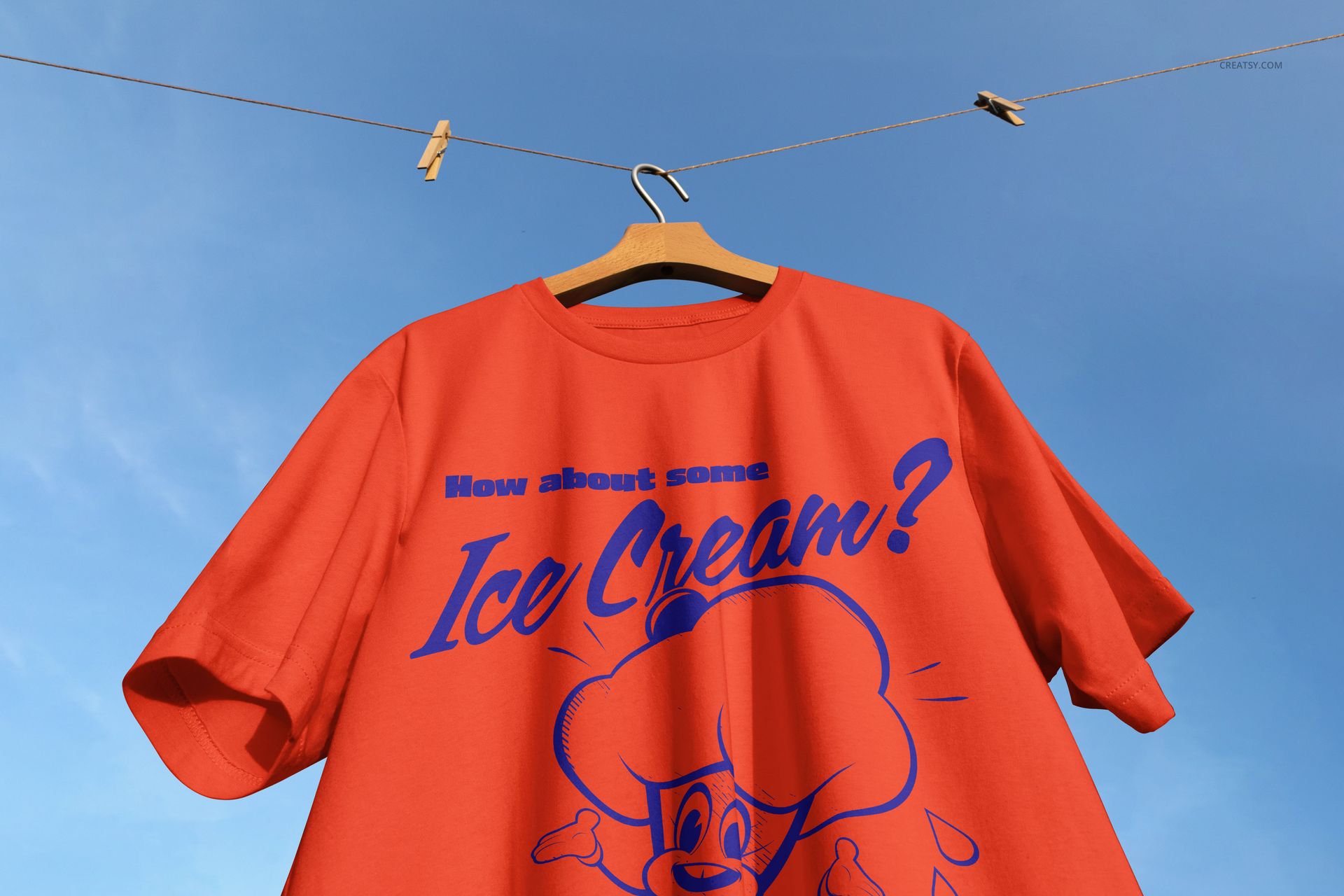 Front view of a red adult t-shirt mockup with blue cartoon graphic and text, hanging on a wooden hanger against a clear sky.
