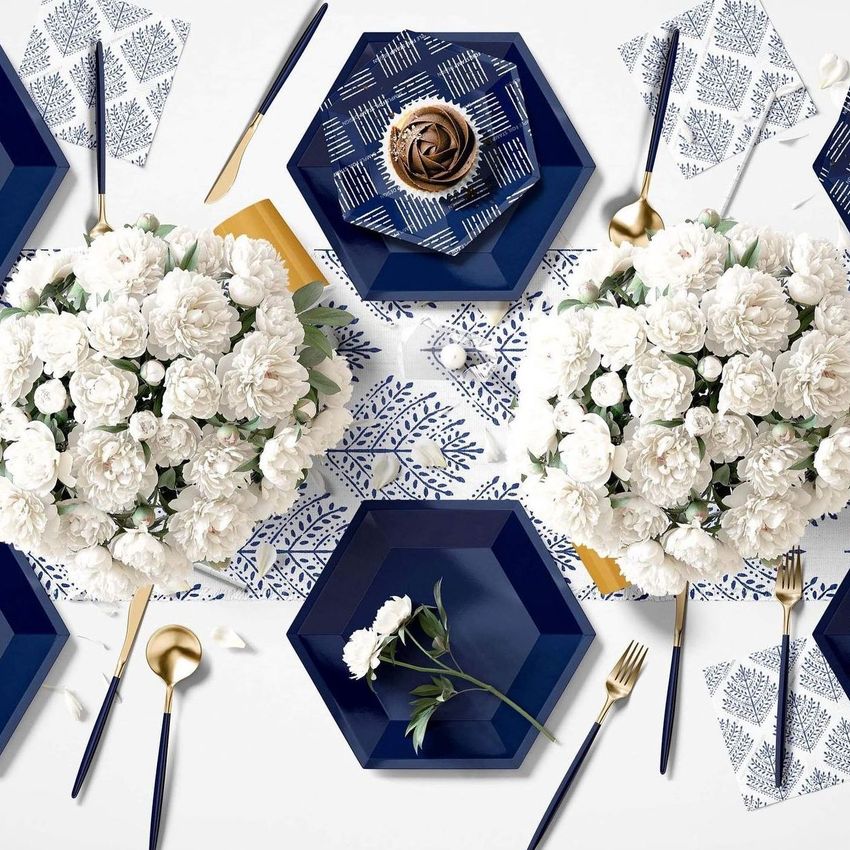 Party supplies mockup featuring navy blue hexagonal plates, gold cutlery, and white floral arrangements on a patterned tablecloth.
