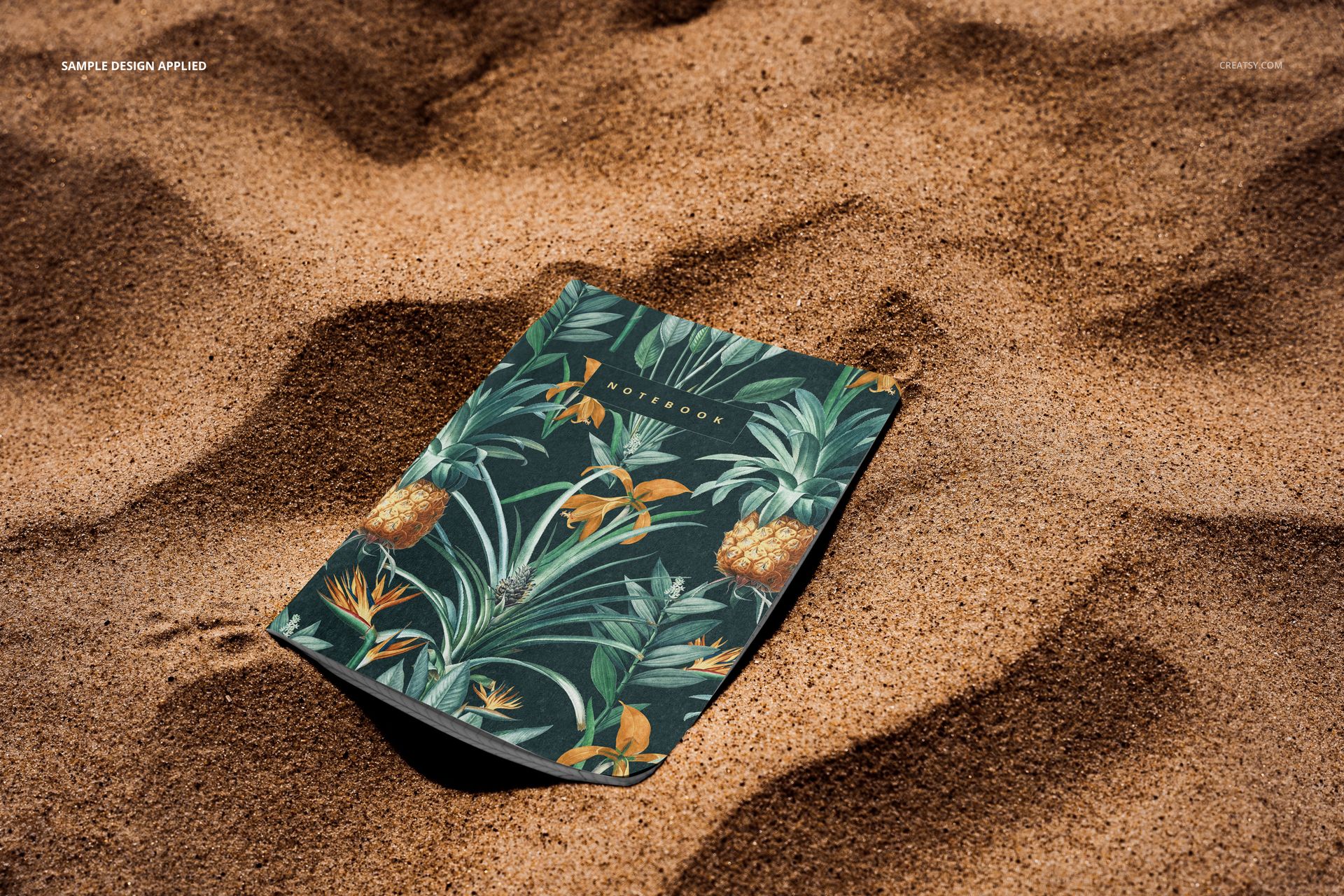PSD mockup of a stationery notebook with a vibrant tropical cover resting on sunlit sand, showing front cover detail.