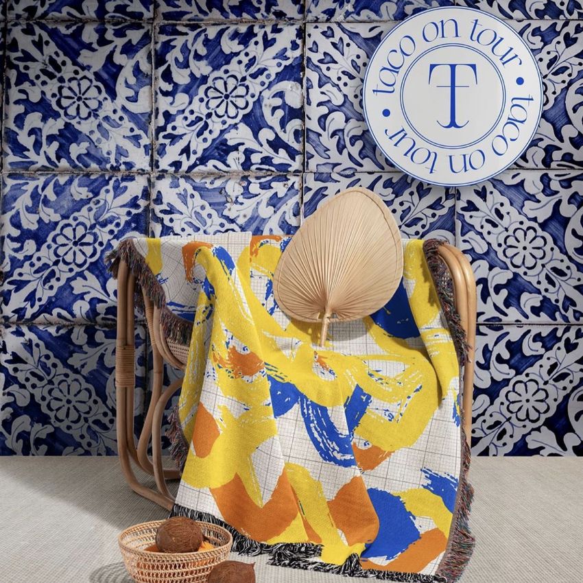 Woven throw blanket mockup draped over a wooden chair, featuring abstract yellow and blue patterns, with a decorative fan and textured tiles in the background.