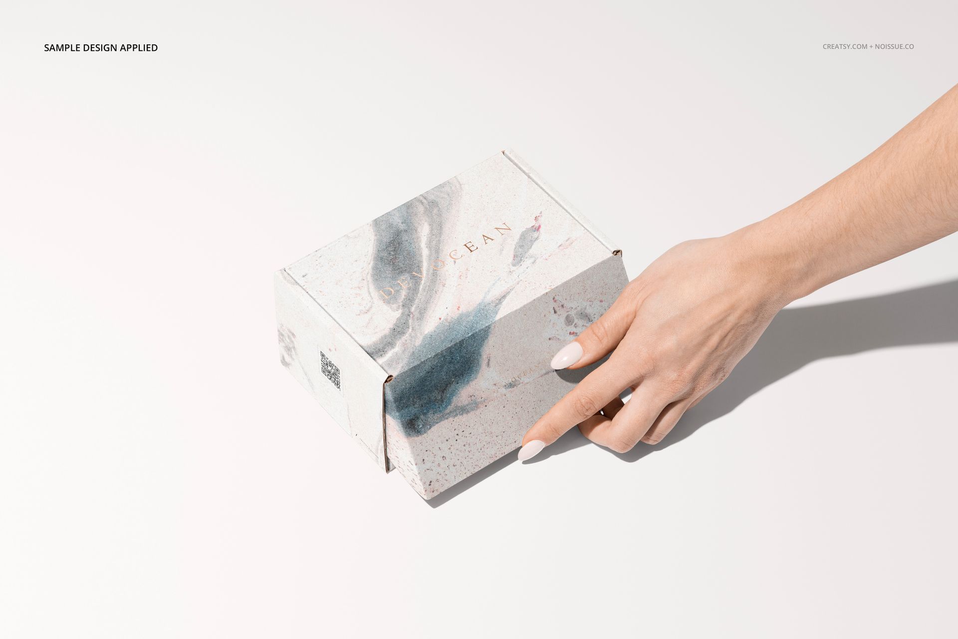 Mockup of a cardboard mailing box featuring abstract blue and pink swirls, viewed from above with a hand on the right.