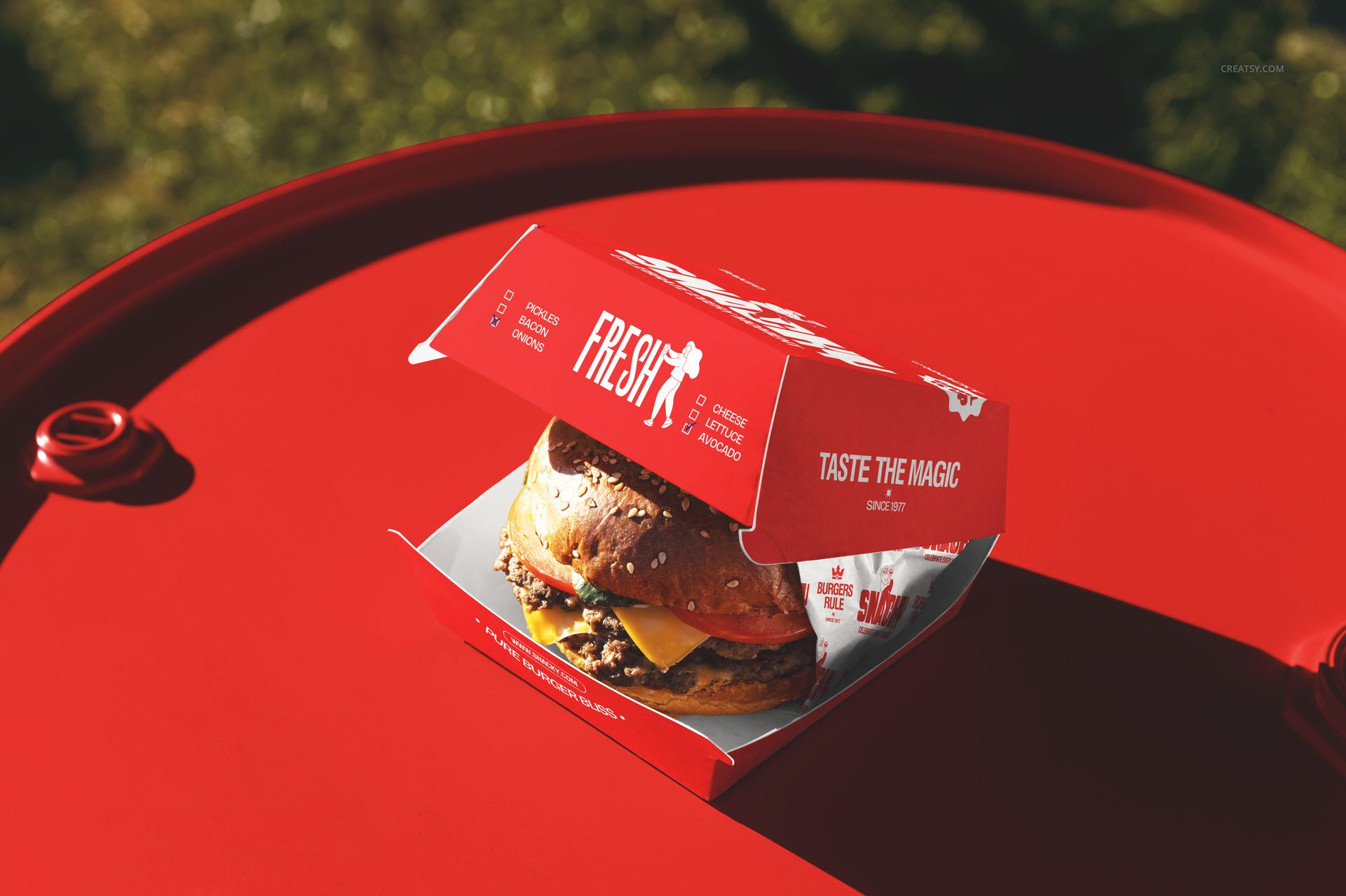 PSD mockup showing a burger with lettuce and cheese in a branded box on a vivid red outdoor table surface.