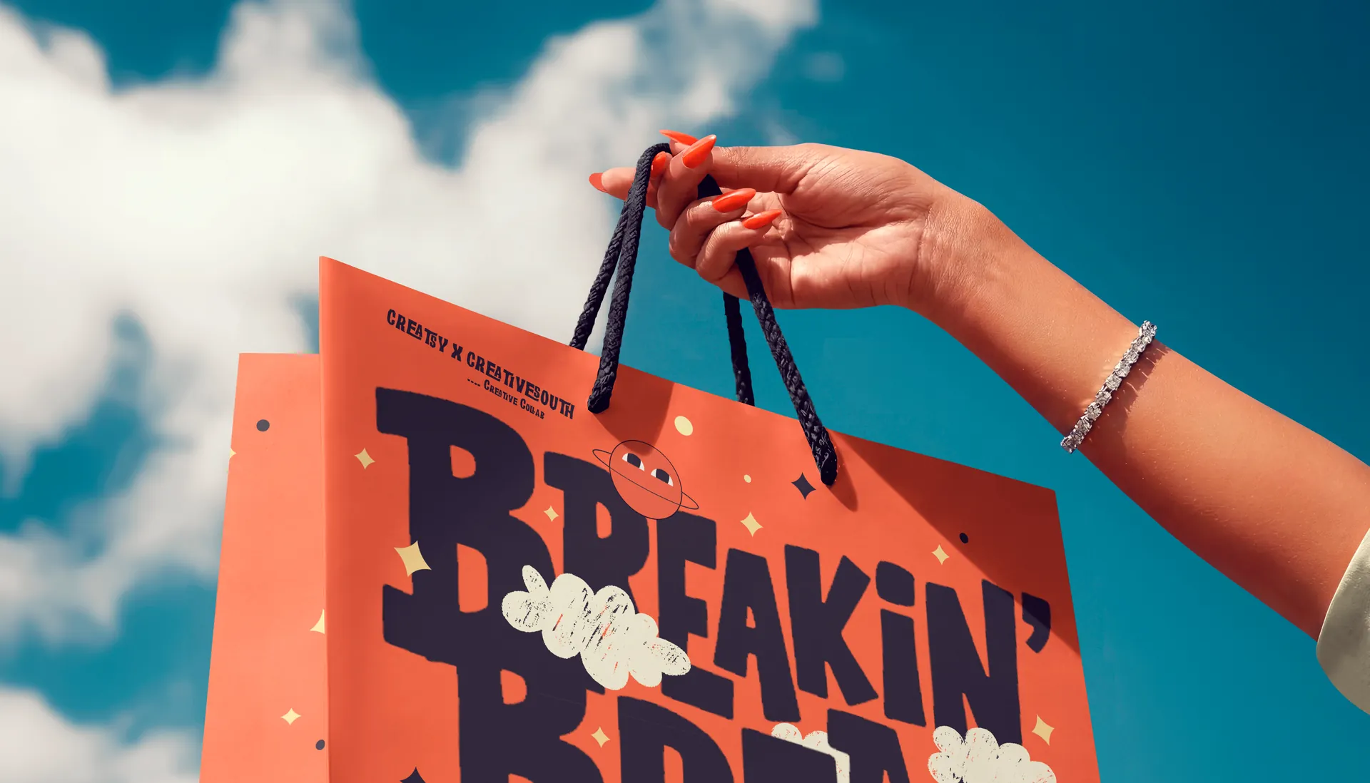 Paper shopping bag mockup with Creative South artwork, held up against a blue sky